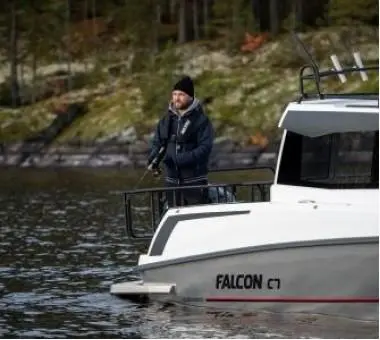 Man in a beanie fishing from a Falcon boat on calm water, surrounded by trees and rocky terrain in a natural setting.